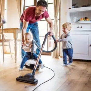 A man and two children cleaning a hardwood floor with a vacuum cleaner.